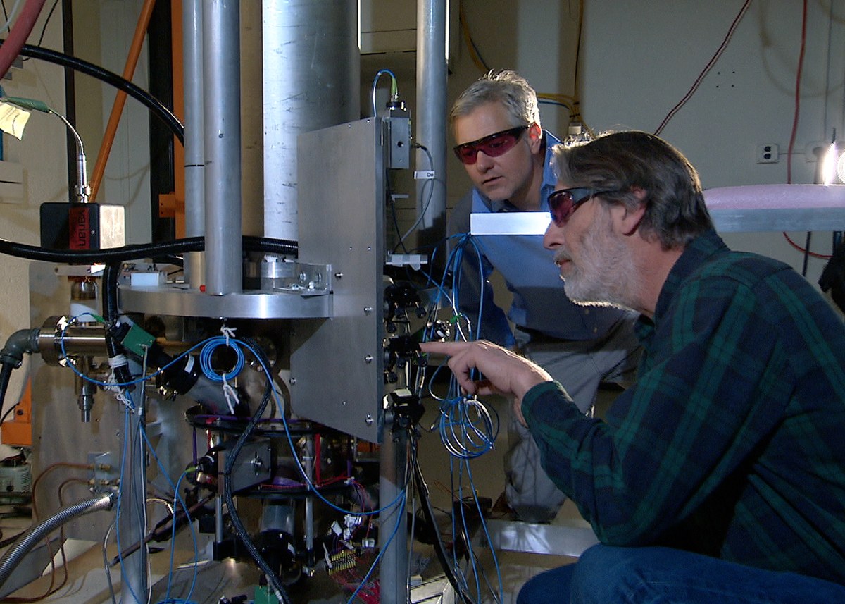 Two NIST physicists beside the NIST-F2 cesium fountain atomic clock, a tall vertical vacuum apparatus surrounded by optics